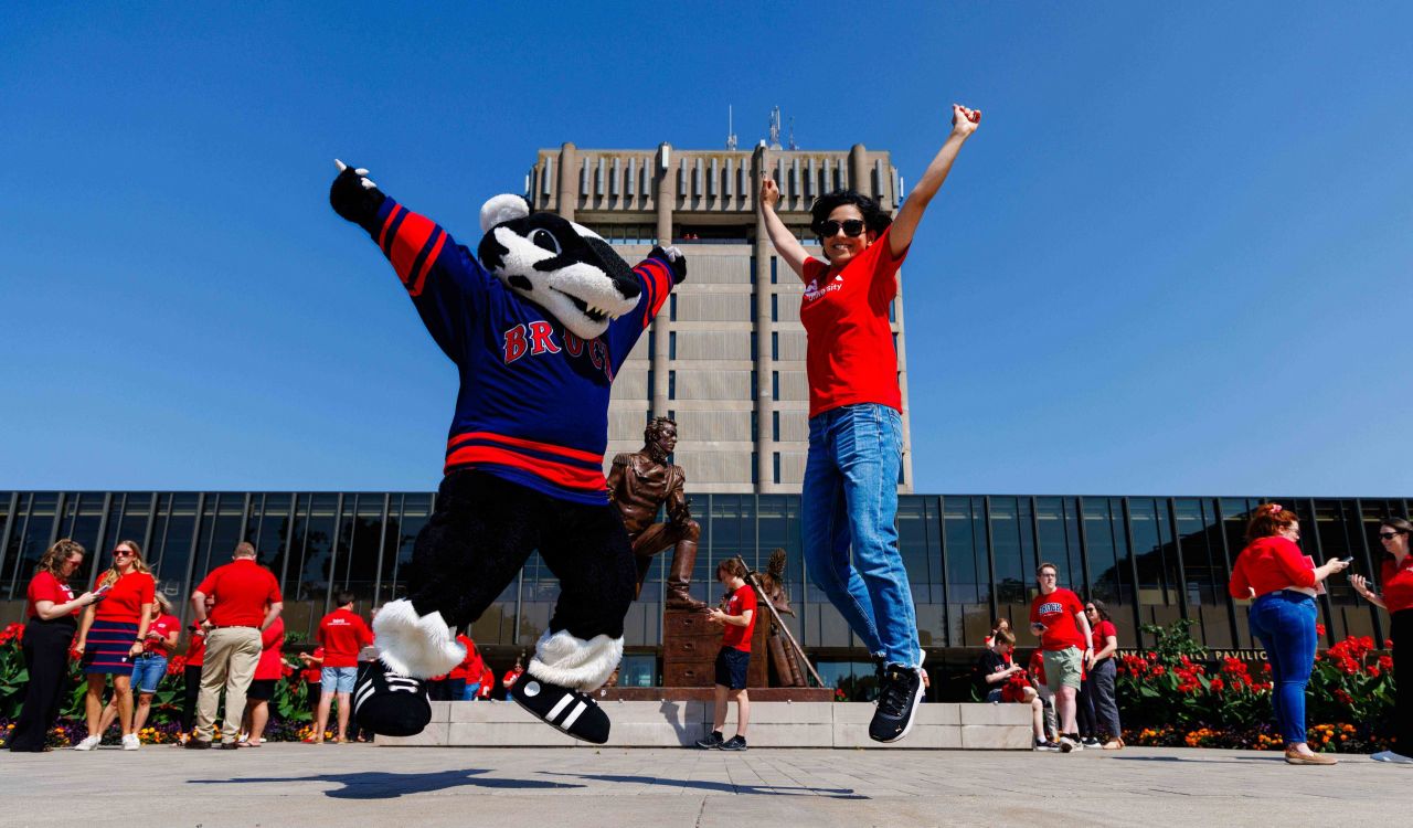 Brock University mascot Boomer the Badger and a student in a red shirt jump with excitement in front of the Schmon Tower during a campus celebration, surrounded by people wearing Brock red.