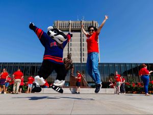 Brock University mascot Boomer the Badger and a student in a red shirt jump with excitement in front of the Schmon Tower during a campus celebration, surrounded by people wearing Brock red.