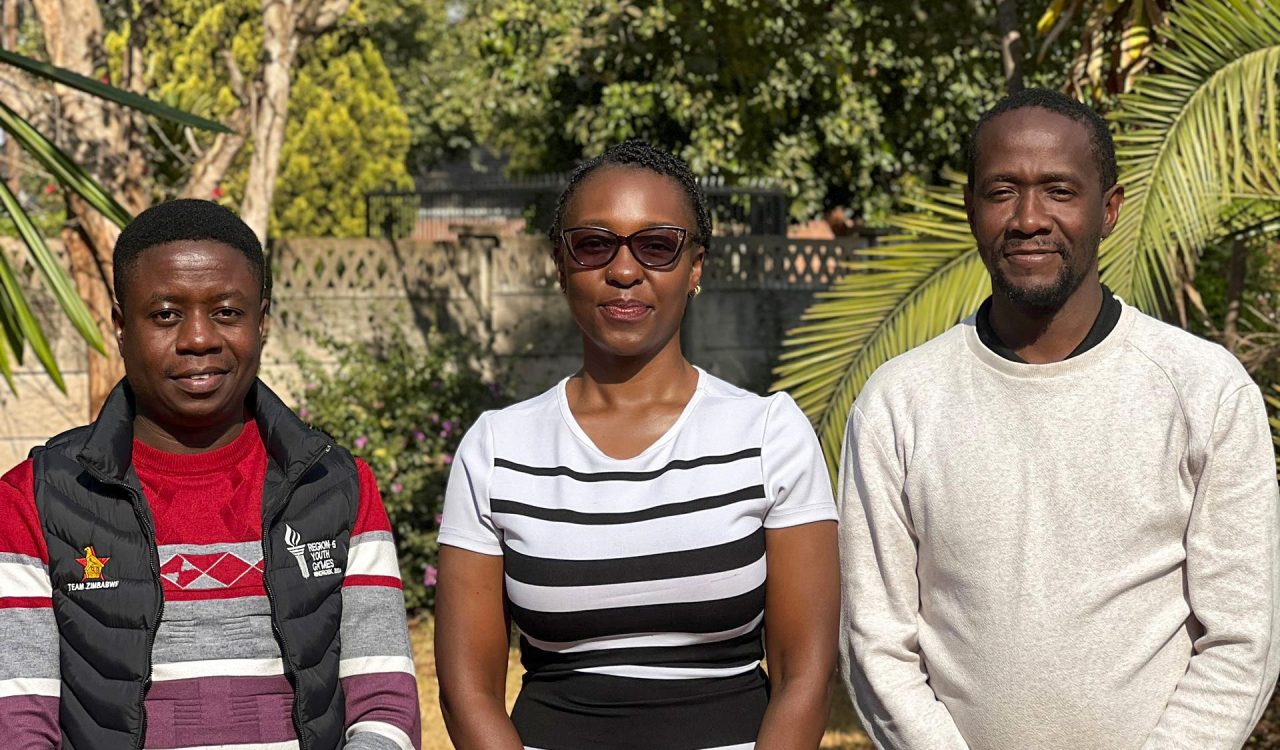 Three people stand together outdoors on a sunny day, posing for a group photo.