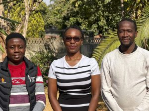 Three people stand together outdoors on a sunny day, posing for a group photo.