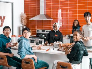 A diverse group of young adults smiling and preparing food together in a bright, modern kitchen with red tiled walls.