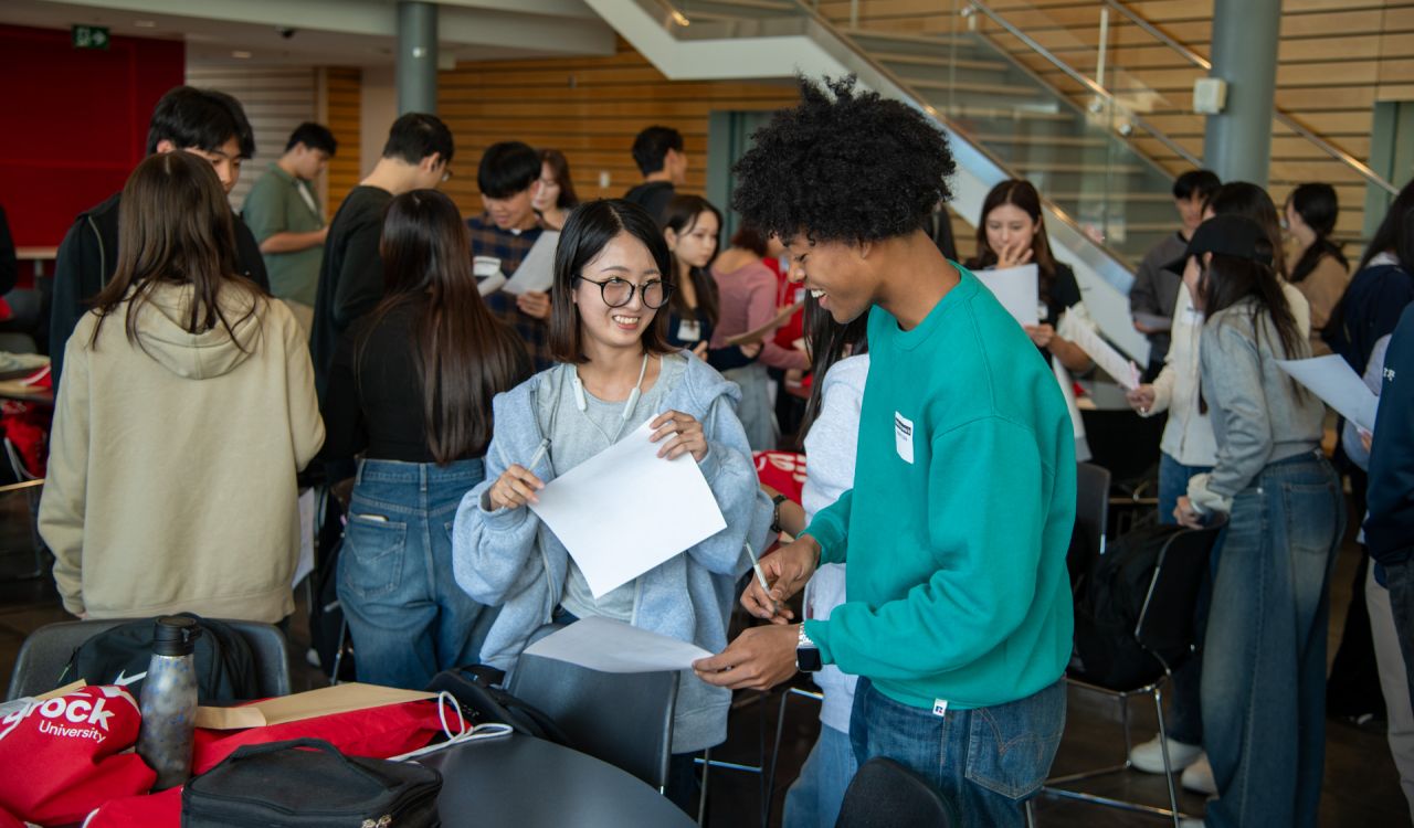 Two students smiling while in conversation in a busy room with crowds in background.