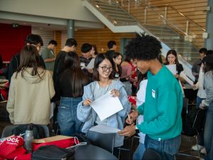 Two students smiling while in conversation in a busy room with crowds in background.
