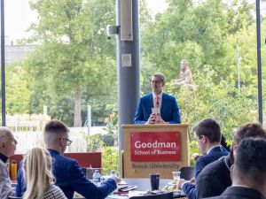 Man in blue suit stands at a podium that says Goodman School of Business Brock University while speaking to people sitting at round tables.