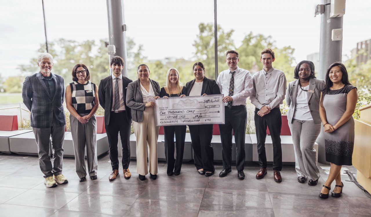 A team of six students receive their prize from four Goodman faculty members, the students hold an oversized cheque that reads $1,000.