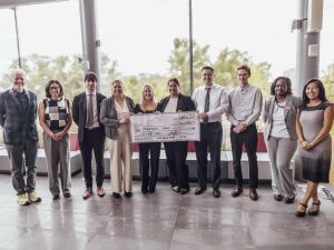 A team of six students receive their prize from four Goodman faculty members, the students hold an oversized cheque that reads $1,000.