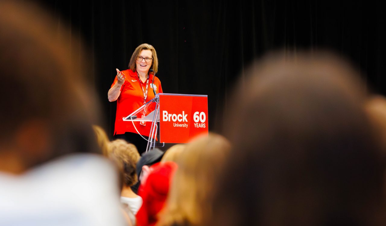A Brock University leader addresses a packed gymnasium during Academic Orientation, speaking from a podium marked with the University’s 60th anniversary logo.
