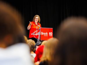 A Brock University leader addresses a packed gymnasium during Academic Orientation, speaking from a podium marked with the University’s 60th anniversary logo.