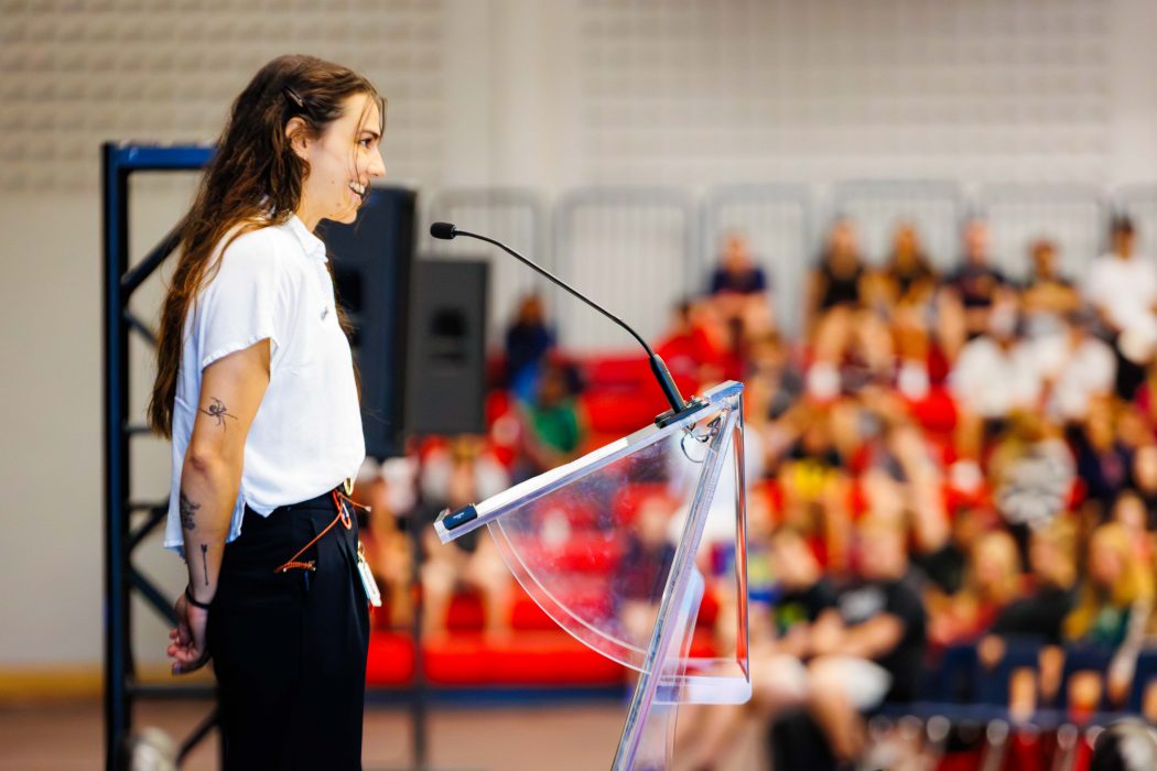 A student leader welcomes peers at Brock University’s Academic Orientation, speaking to the crowd from a clear podium inside the gymnasium.