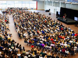 Hundreds of students gather in a large campus gymnasium for an academic orientation session, listening to speakers on stage as part of the welcome programming.