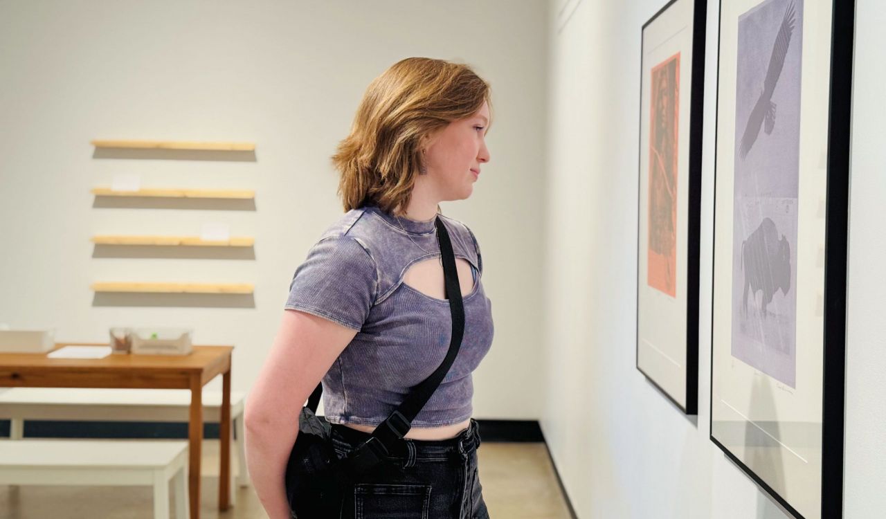 A woman stands in an art gallery looking at a painting. A table and chairs can be seen in the background.