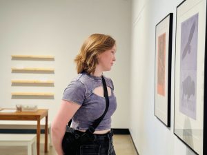 A woman stands in an art gallery looking at a painting. A table and chairs can be seen in the background.