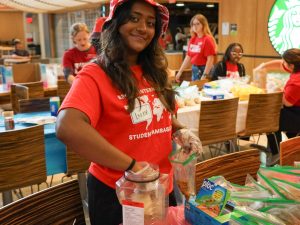 A university student in a red shirt and hat packs food at a volunteer event.