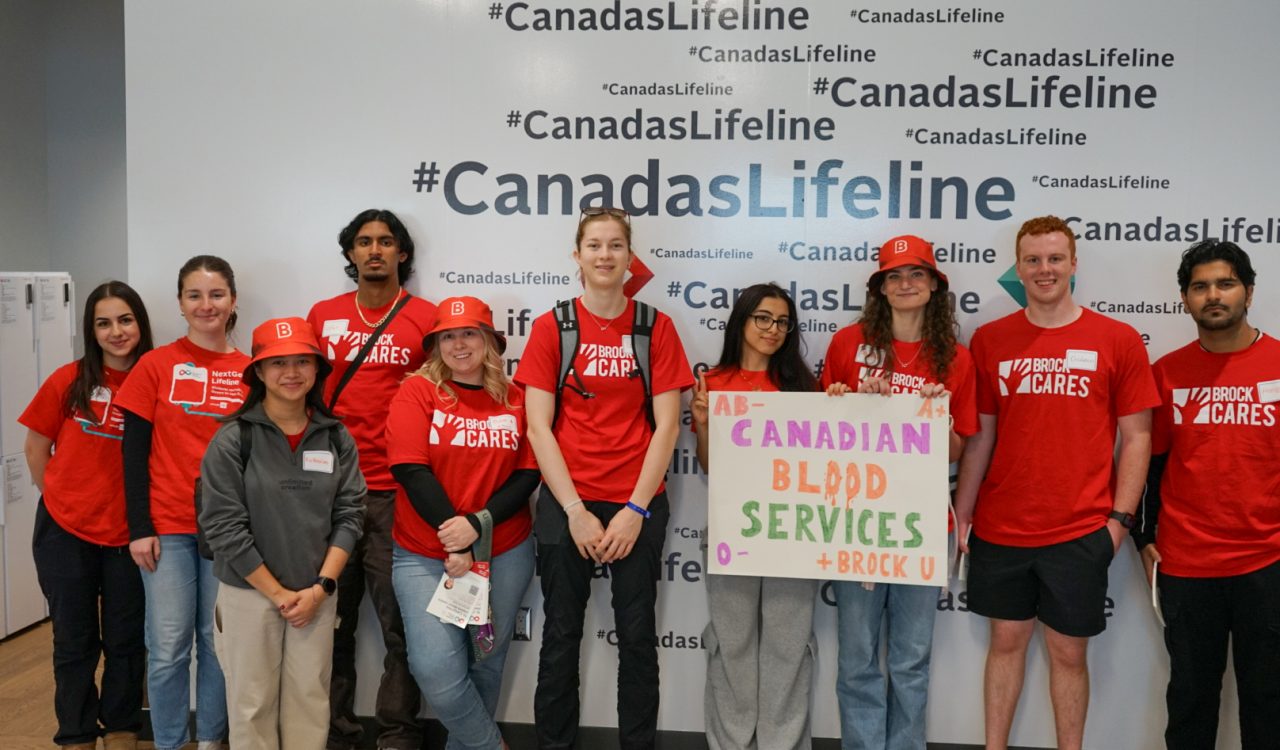 A group of university students in red shirts pose with a "Canadian Blood Services" sign at a volunteering event.