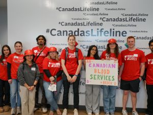 A group of university students in red shirts pose with a "Canadian Blood Services" sign at a volunteering event.