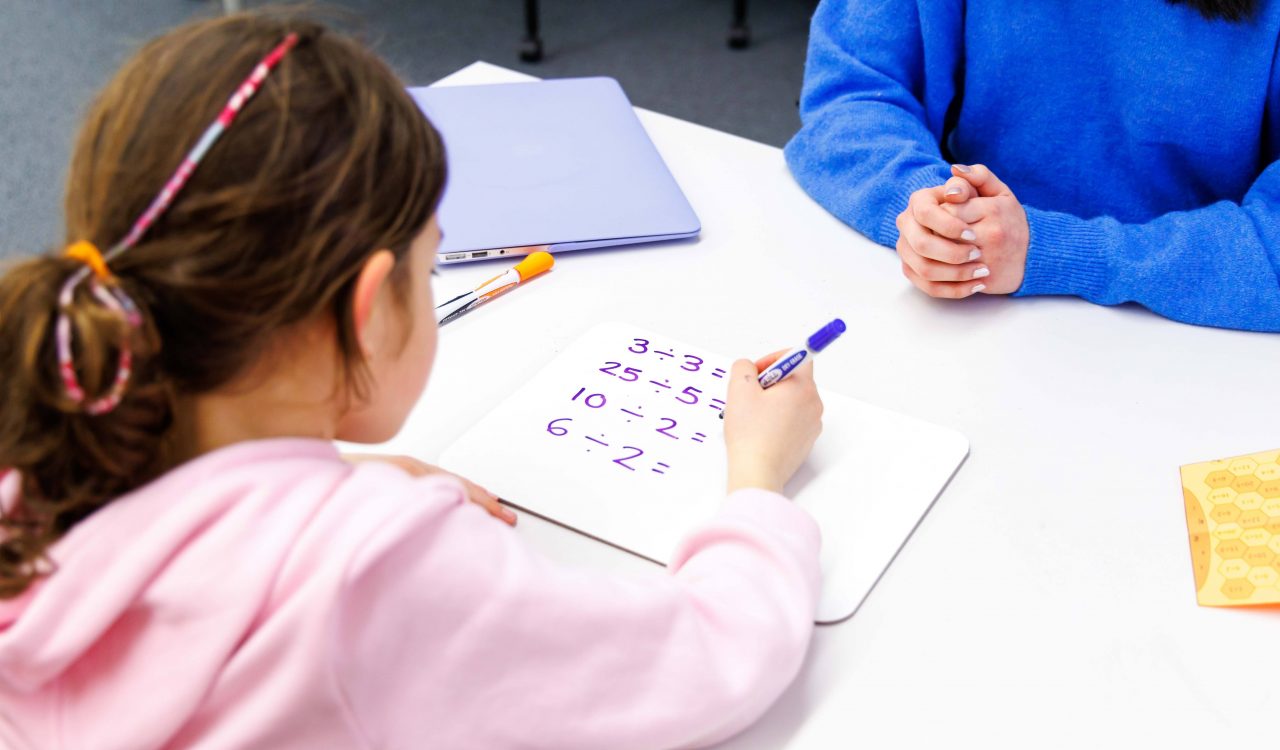 A young girl is seen over the shoulder, sitting at a white desk doing math equations while sitting across from a tutor.