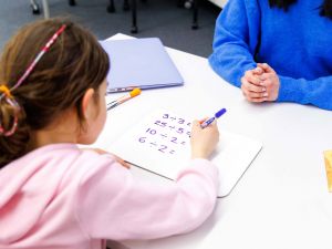A young girl is seen over the shoulder, sitting at a white desk doing math equations while sitting across from a tutor.