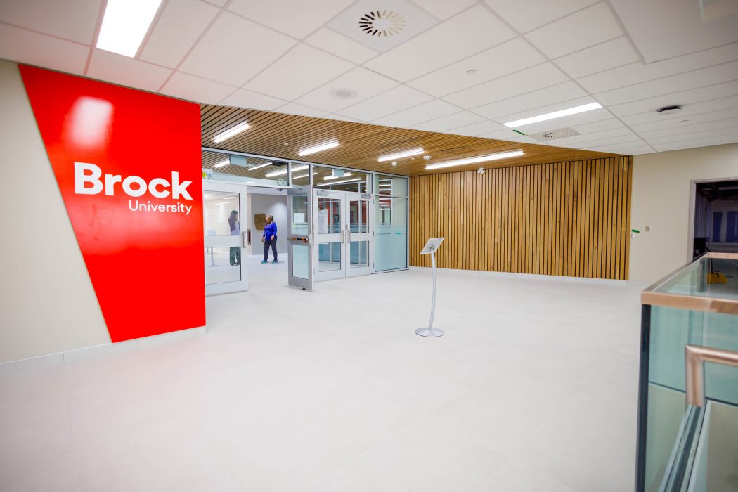 The newly renovated entrance space at Brock University’s Burlington campus, featuring a bold red wall with the Brock University logo, modern wood paneling, and glass doors leading into the campus.