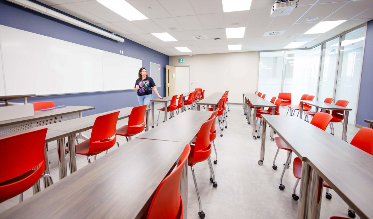 Bright, modern classroom with rows of red chairs and long desks, featuring a large whiteboard and ceiling-mounted projector. A person stands near the front of the room by the whiteboard.