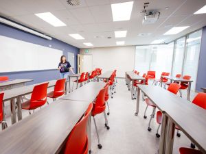 Bright, modern classroom with rows of red chairs and long desks, featuring a large whiteboard and ceiling-mounted projector. A person stands near the front of the room by the whiteboard.