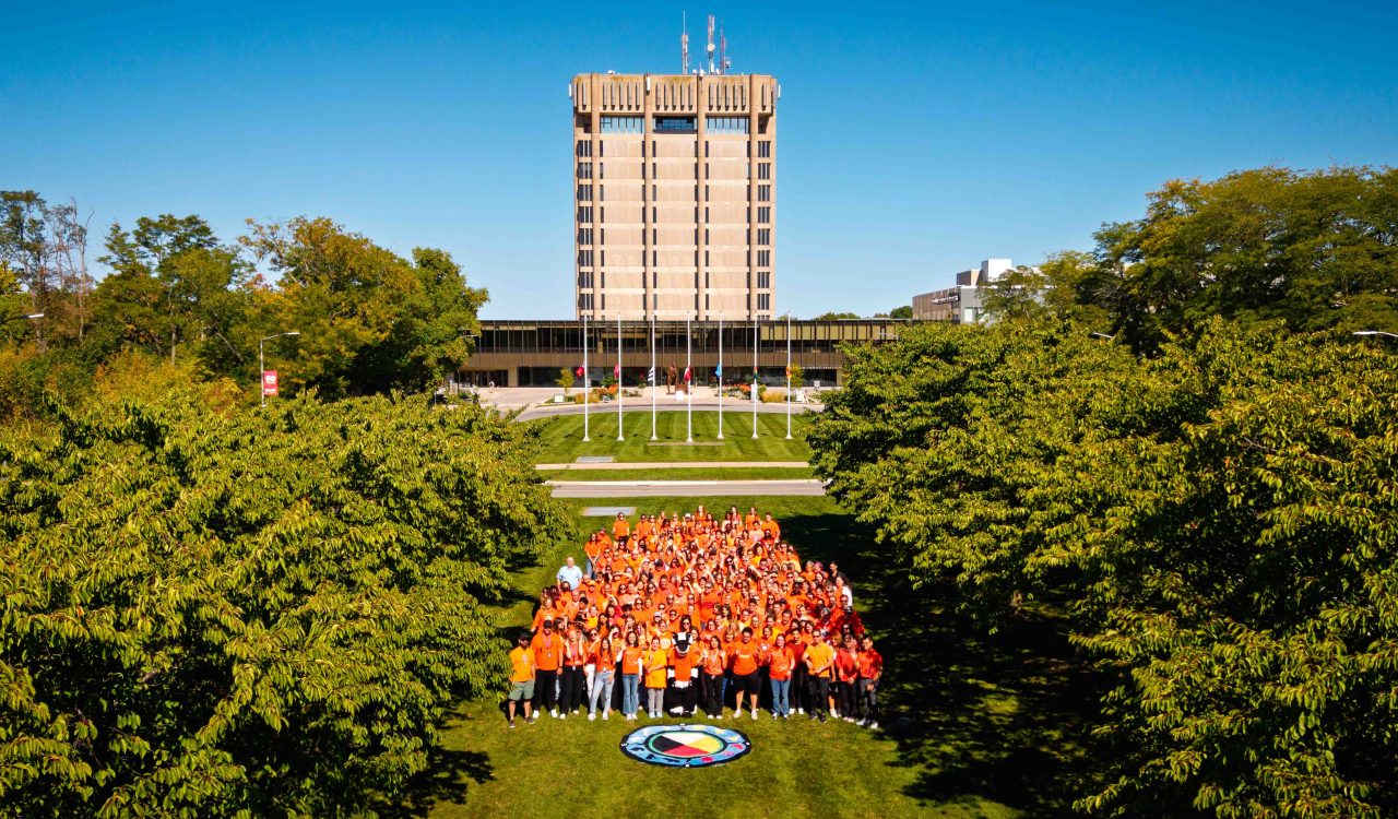 A large group of people wearing orange shirts gathered on a grassy lawn in front of a tall concrete campus tower building on a clear sunny day, with green trees framing the scene.