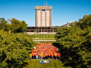 A large group of people wearing orange shirts gathered on a grassy lawn in front of a tall concrete campus tower building on a clear sunny day, with green trees framing the scene.