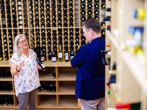 Woman holding a wine bottle speaks to a man inside a wine cellar.