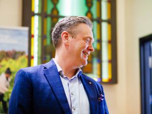 Man in blue blazer smiling indoors near a stained-glass window.