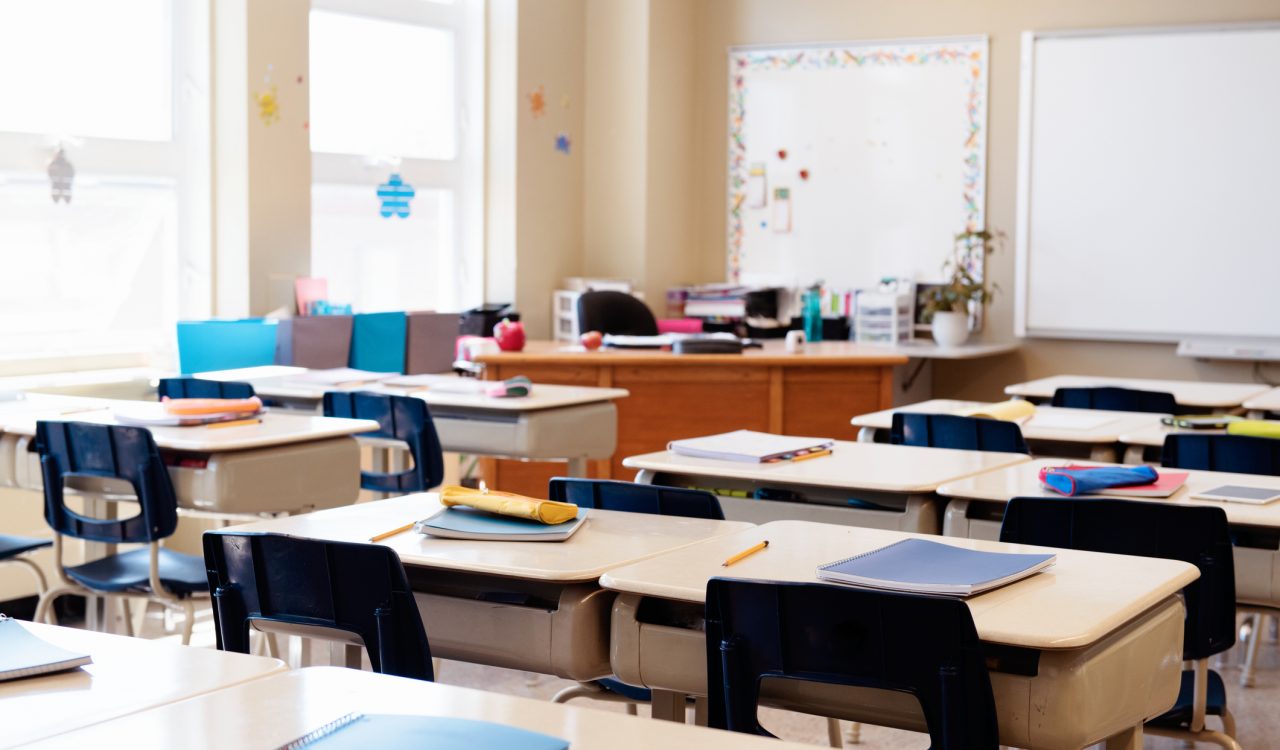Rows of desks holding school supplies in an empty, sunny classroom.