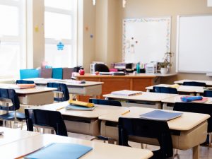 Rows of desks holding school supplies in an empty, sunny classroom.
