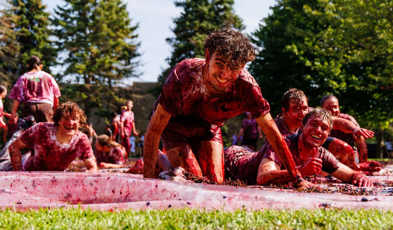 Students covered in smashed grapes crawl and laugh across a tarp during a grape stomp event on a sunny day.