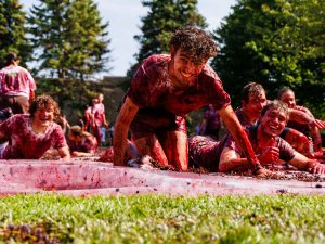 Students covered in smashed grapes crawl and laugh across a tarp during a grape stomp event on a sunny day.
