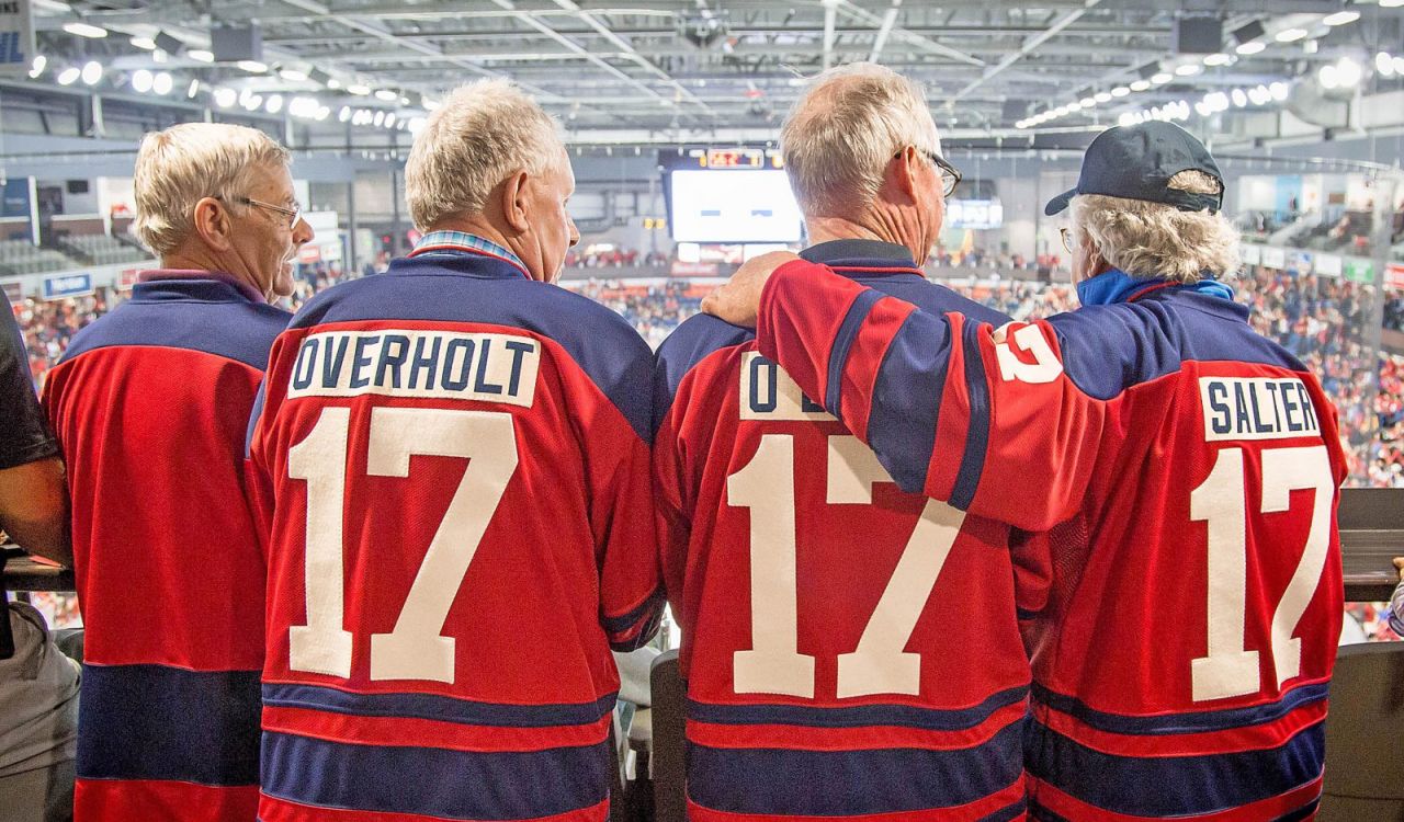 Four hockey fans wearing matching red and blue jerseys with the number 17 and their names on the back stand together in an arena, watching the game.