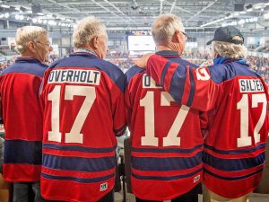 Four hockey fans wearing matching red and blue jerseys with the number 17 and their names on the back stand together in an arena, watching the game.