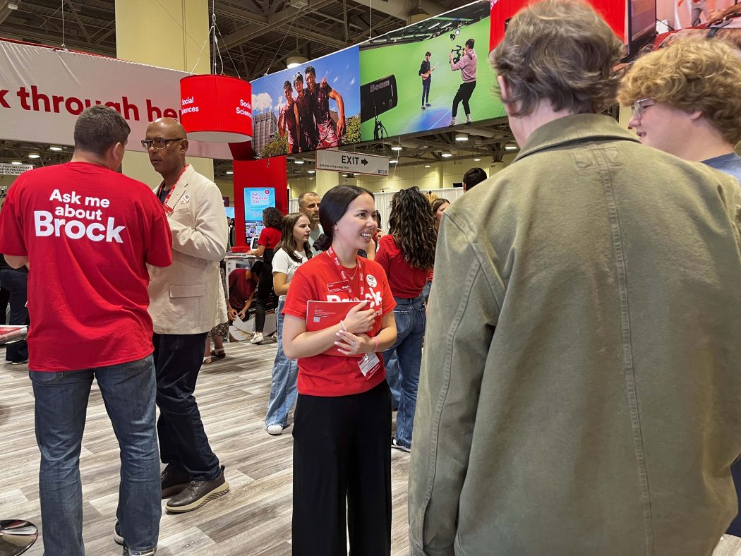 A young woman in a red shirt speaks to a family at a university information fair.