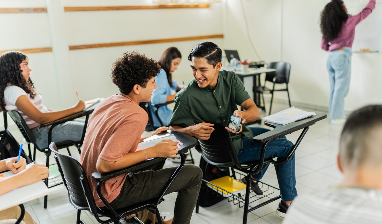 Two teenage boys laugh and sneak a look at a video on a cellphone while in class.