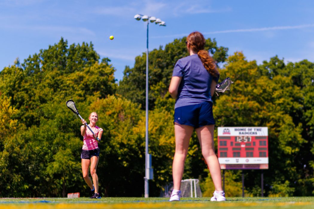 Two young women practice lacrosse on a sunny field.