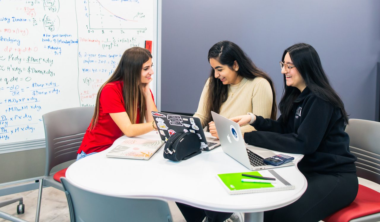 Three students sit around a white table with laptops, notebooks and a calculator, smiling and collaborating in a study session.
