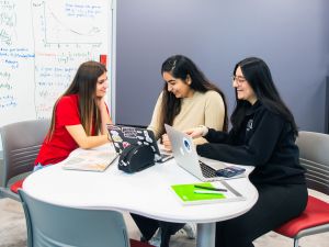 Three students sit around a white table with laptops, notebooks and a calculator, smiling and collaborating in a study session.