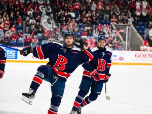 Brock Badgers men's hockey players on ice in a packed arena. One player lifts his glove to his ear, encouraging the crowd to cheer louder.