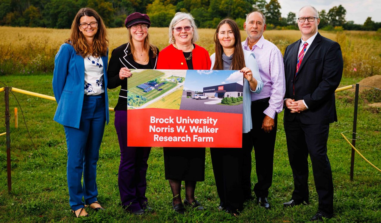 A group of six people pose with a poster board for the Brock University Norris W. Walker Research Farm in a field.