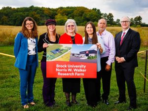 A group of six people pose with a poster board for the Brock University Norris W. Walker Research Farm in a field.