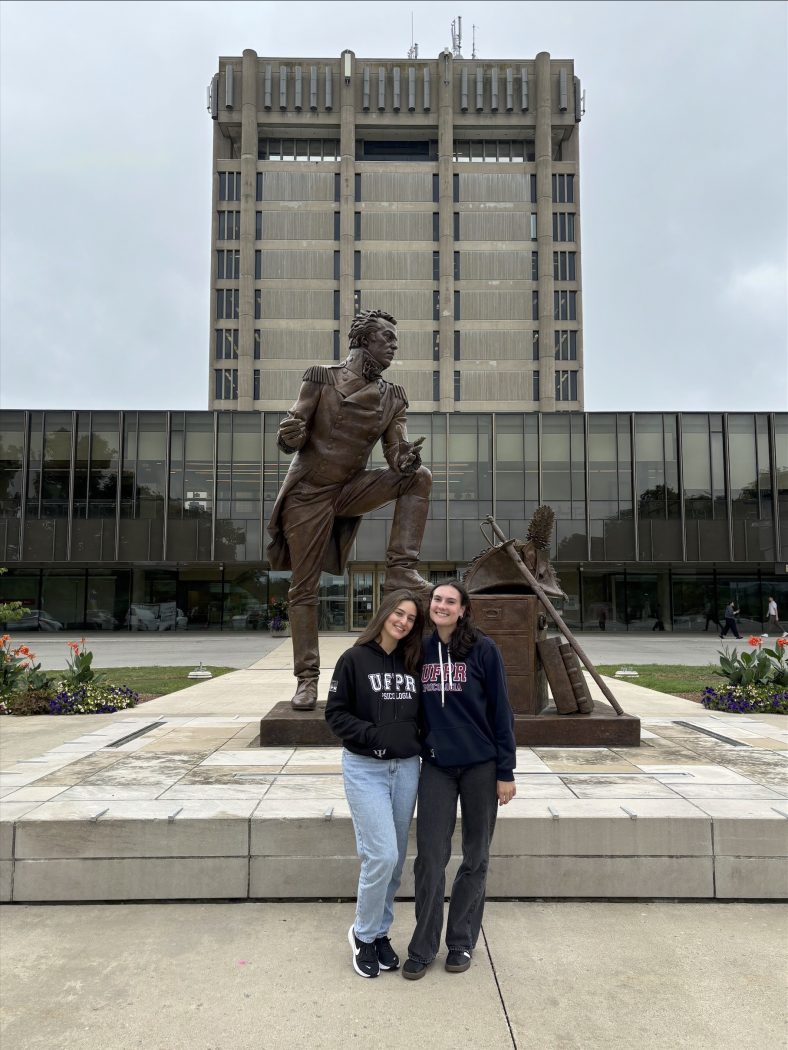 Two university students stand in front of a statue outside a concrete tower.