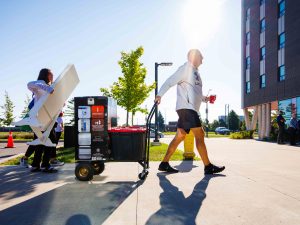 Two people carry a student's belongings into a university residence building.