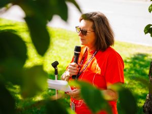 Woman wearing orange shirt speaks to group of people.