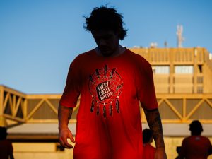 A lacrosse player in an orange ‘Every Child Matters’ T-shirt stands in shadow against a blue sky.
