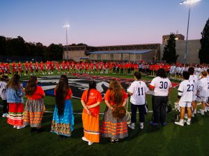 Players and community members form a large circle at midfield during a sunset ceremony.
