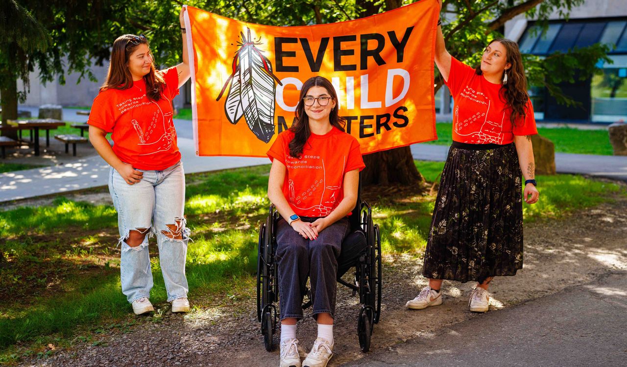 Three women wearing bright orange T-shirts with the words ‘Taking Steps Towards Reconciliation’ stand together outdoors.Three women wearing bright orange T-shirts with the words ‘Taking Steps Towards Reconciliation’ stand together outdoors.