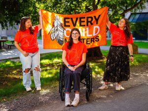 Three women wearing bright orange T-shirts with the words ‘Taking Steps Towards Reconciliation’ stand together outdoors.Three women wearing bright orange T-shirts with the words ‘Taking Steps Towards Reconciliation’ stand together outdoors.