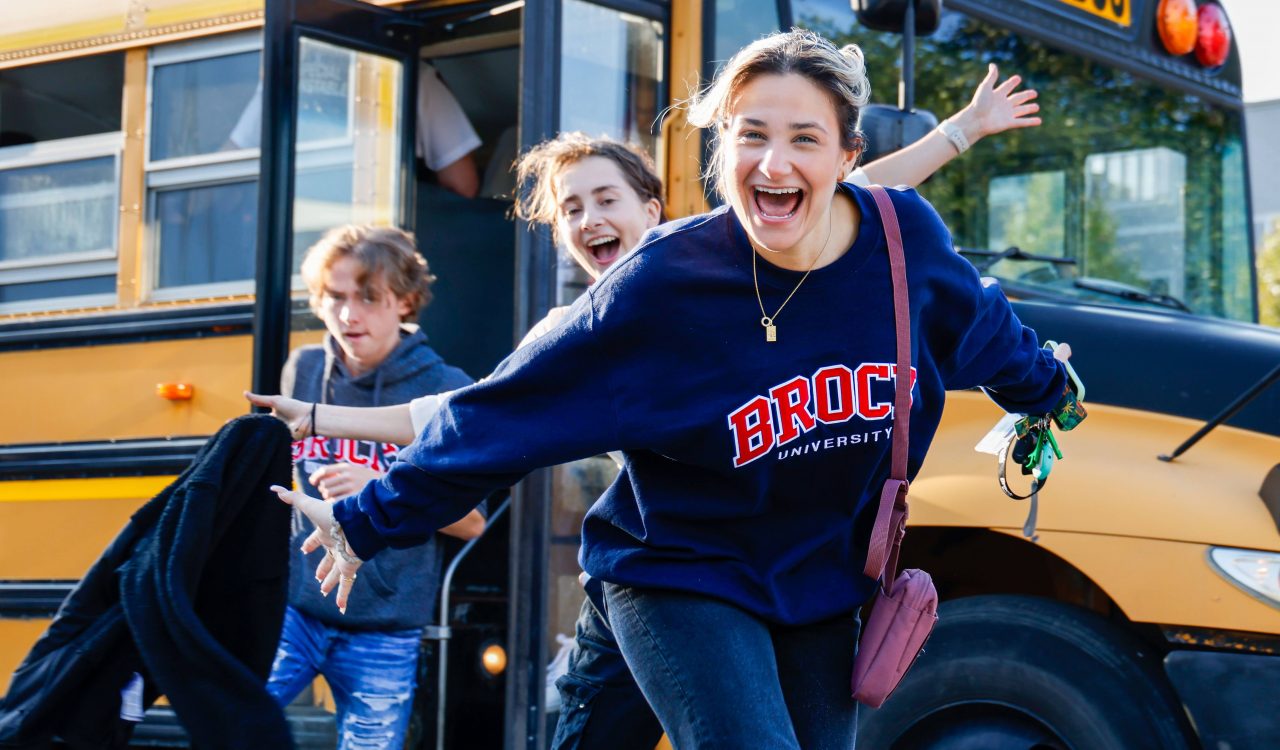 Three students, one smiling broadly in a Brock University sweatshirt, step off a yellow school bus, as they arrive for an off-campus event.